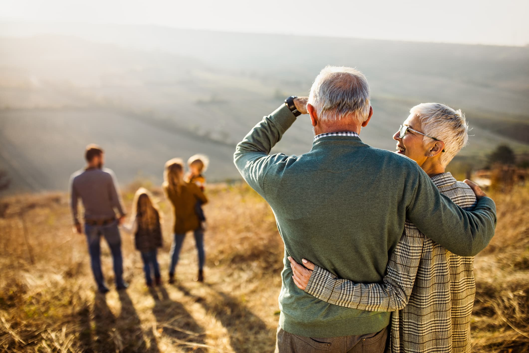 Grandparents standing together on a hillside at sunset, watching their children and grandchildren walk ahead of them.